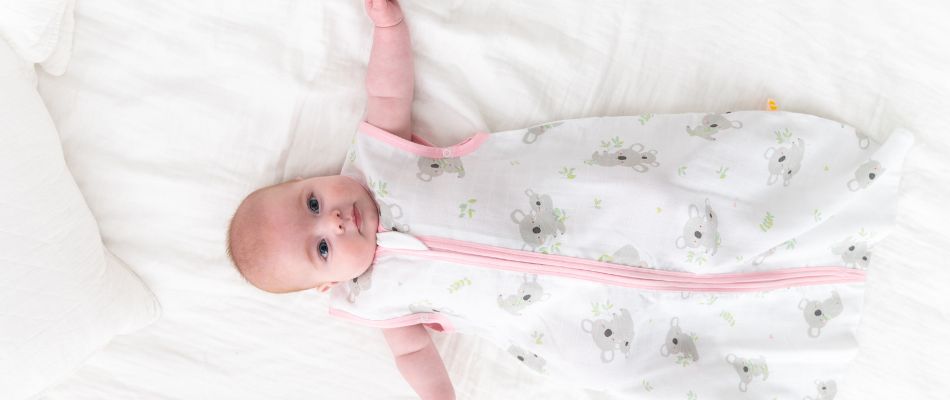 Ein Baby liegt in einem weißen Schlafsack mit grauen Koala-Motiven und einem rosa Reißverschluss auf einem weißen Bett.