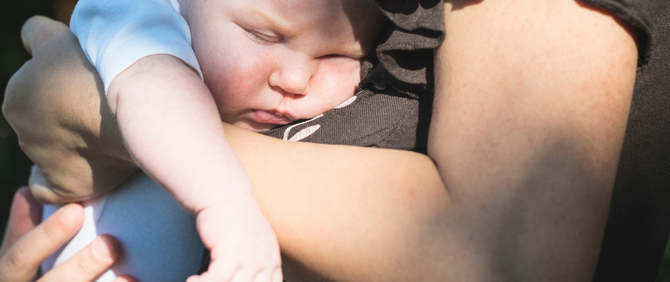 Ein Baby schläft in den Armen einer Person, die dunkle Kleidung trägt. Das Baby trägt einen weißen Strampler und ist festgehalten und getröstet.