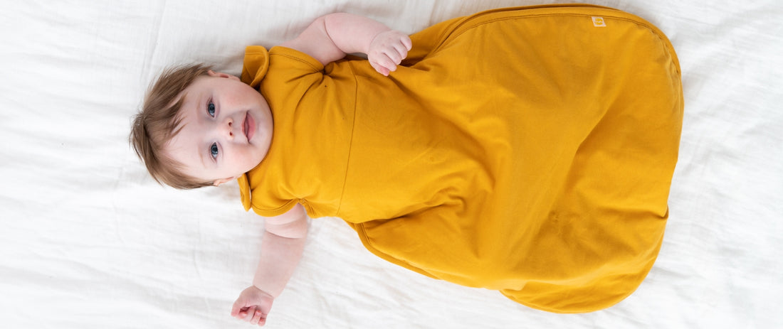 Ein Baby liegt auf dem Rücken auf einem weißen Bett und trägt einen gelben Schlafsack. Es schaut direkt in die Kamera und wirkt entspannt und zufrieden.