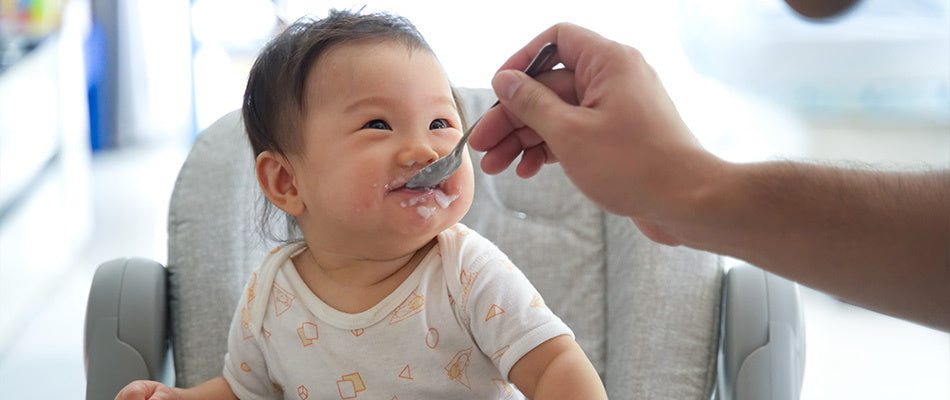 Ein Baby sitzt in einem Hochstuhl und lächelt, während es mit einem Löffel gefüttert wird. Das Baby trägt einen hellen Strampler mit einem Muster, und es hat etwas Nahrung um den Mund.