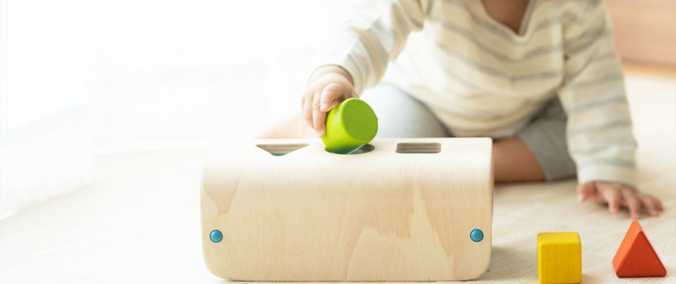 Ein Baby spielt mit einem Holzspielzeug, in das verschiedene Formen passen. Es hält eine grüne Form in der Hand.