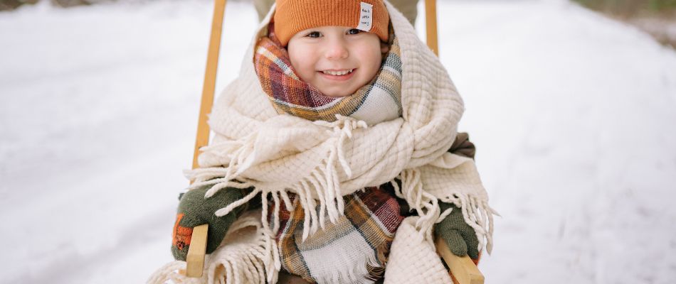 Kind in Winterkleidung sitzt auf einem Schlitten, umgeben von Schnee und lächelt.