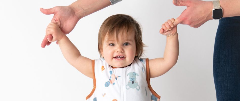 Ein Baby lächelt fröhlich, während es von zwei Erwachsenen an den Händen gehalten wird, die ihm beim Laufenlernen helfen.