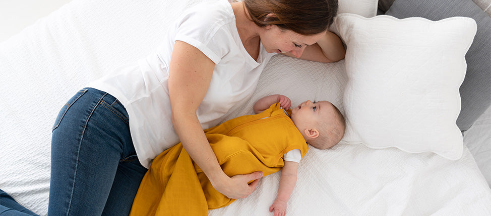 Eine Mutter liegt auf einem Bett und schaut liebevoll ihr Baby an, das in einem gelben Schlafsack neben ihr liegt. Die Mutter trägt ein weißes T-Shirt und blaue Jeans, während das Baby in dem Schlafsack auf einer weißen Bettdecke liegt.