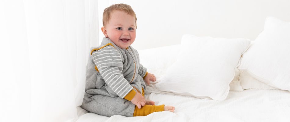 Lächelndes Baby sitzt selbstständig auf einem weißen Bett in einem grauen Schlafanzug mit gelben Bündchen.