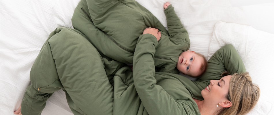 Eine Mutter und ihr Baby liegen nebeneinander auf einem Bett. Beide tragen passende grüne Schlafsäcke  und lächeln. Die Mutter hat ihren Arm um das Baby gelegt, das sie liebevoll ansieht.