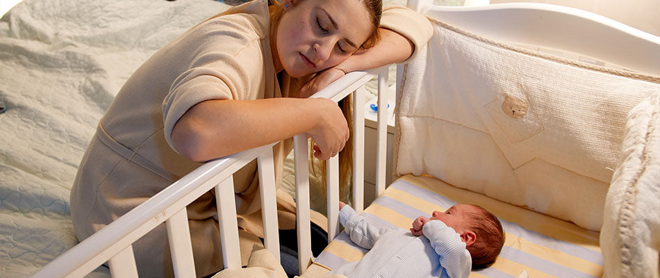 Eine erschöpfte Mutter lehnt sich auf das Gitterbett, während ihr Baby darin liegt und wach ist.