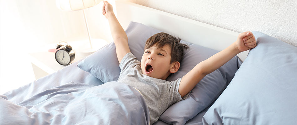 Ein Junge, der in seinem Bett liegt und sich am Morgen streckt, während er gähnt. Im Hintergrund ist ein Wecker auf dem Nachttisch zu sehen.