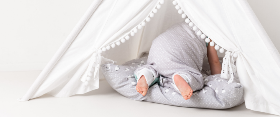 Ein Baby krabbelt in ein weißes Zelt mit Pompon-Dekorationen. Es trägt einen grauen Strampler mit weißen Punkten und liegt auf einem grauen Kissen mit weißen Mustern.