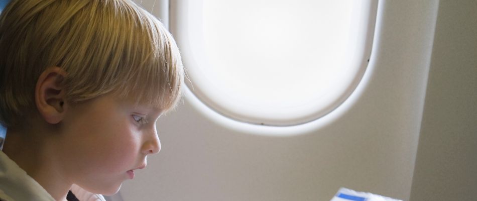 Ein blondes Kind sitzt im Flugzeug und schaut konzentriert auf etwas in seinen Händen. Im Hintergrund ist das Fenster des Flugzeugs zu sehen