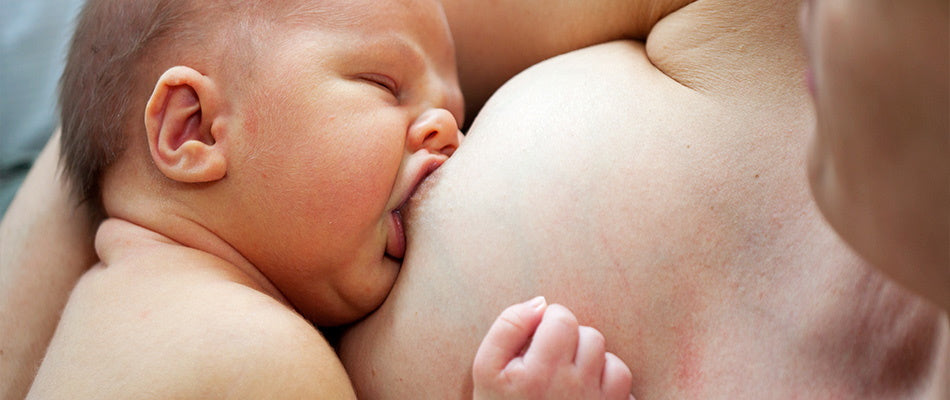 Ein Baby trinkt an der Brust seiner Mutter. Das Baby hat die Augen geschlossen und wirkt entspannt. Es hält die Brust der Mutter mit einer Hand und ist dabei, sich zu ernähren.