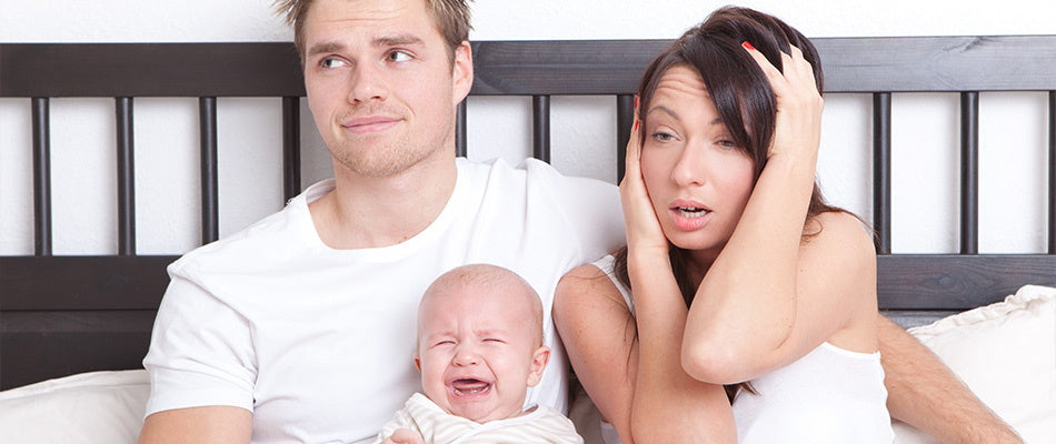 Eine müde aussehende Mutter und ein erschöpfter Vater sitzen auf einem Bett, während ein Baby zwischen ihnen weint.