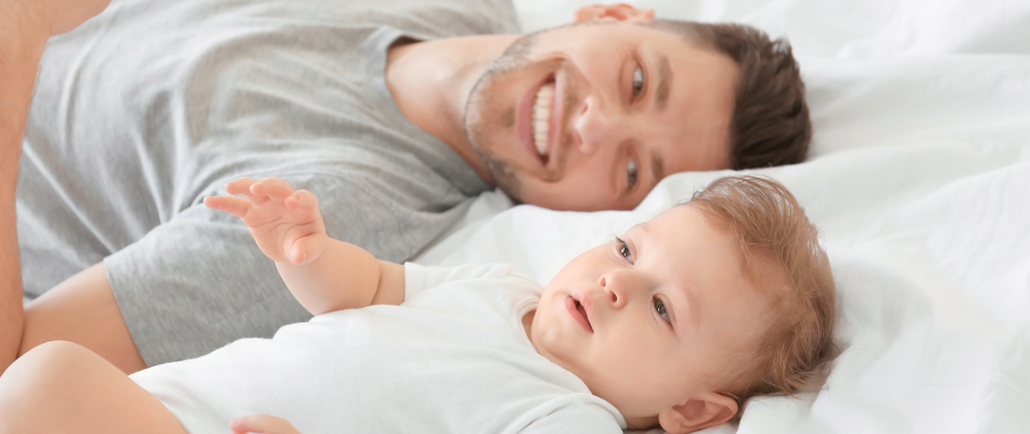 Ein lächelnder Mann liegt auf dem Bett neben einem Baby, das ebenfalls auf dem Bett liegt. Beide tragen helle Kleidung.