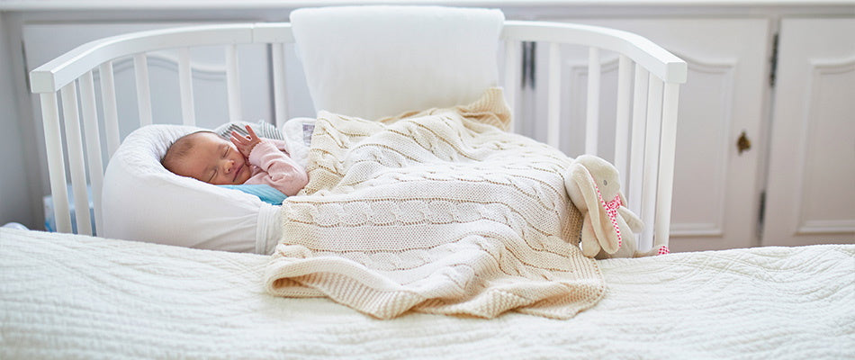 Baby schläft friedlich in einem weißen Gitterbett, das direkt an das Elternbett angeschlossen ist. Das Baby ist in eine beige Strickdecke eingewickelt.