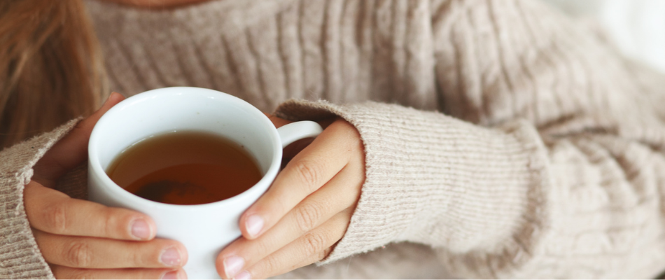 Eine Person in einem beigen Strickpullover hält eine weiße Tasse mit Tee in beiden Händen.