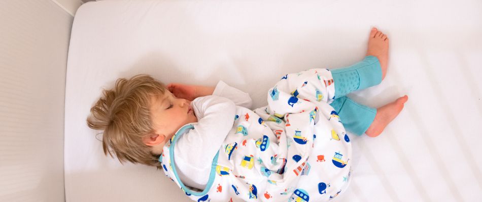 Ein kleines Kind schläft friedlich in einem Bett. Es trägt einen bunten Schlafsack mit Füßen