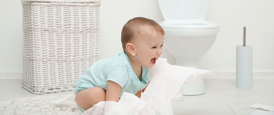 Ein Baby sitzt lachend auf dem Boden eines Badezimmers und spielt mit Toilettenpapier. Im Hintergrund sind eine Toilette, eine Toilettenpapierrolle und ein weißer Wäschekorb zu sehen.