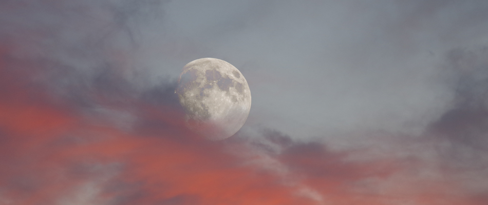 Ein Vollmond ist am Himmel zu sehen, umgeben von rot und violett gefärbten Wolken während der Dämmerung.