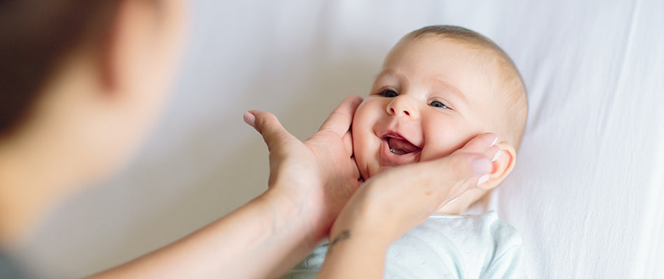 Elternteil hält die Wangen eines lachenden Babys, das auf dem Rücken liegt.