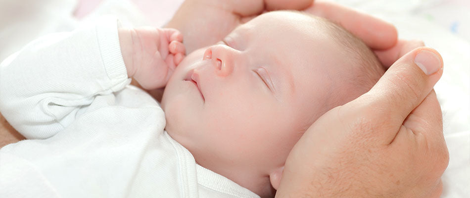 Ein schlafendes Baby in einem weißen Strampler wird liebevoll von zwei Händen am Kopf gehalten.