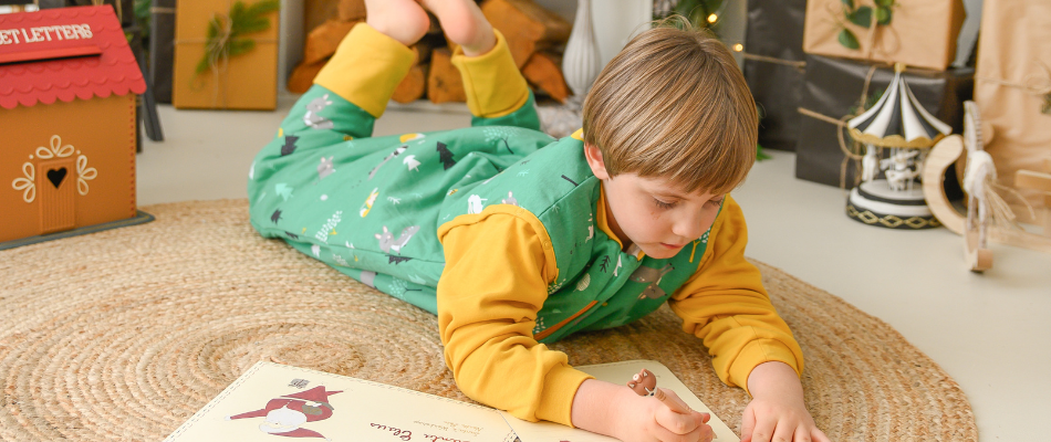 Ein Kind liegt auf einem runden, geflochtenen Teppich und malt in einem Buch. Es trägt einen grünen Schlafanzug mit Waldmotiven und gelben Ärmeln. Im Hintergrund sind weihnachtliche Dekorationen zu sehen.