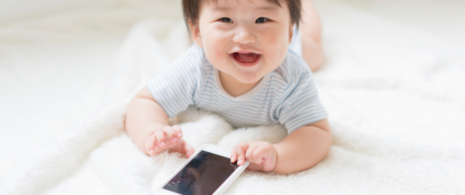 Ein Baby liegt auf dem Bauch auf einer weichen, weißen Decke und lächelt fröhlich in die Kamera. Vor ihm liegt ein Smartphone