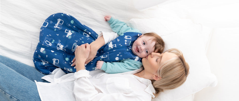 Eine Mutter liegt auf einem Bett und hält ihr Baby, das in einem blauen Schlafsack mit Fahrzeugmotiven steckt. Sie küsst das Baby liebevoll auf die Wange.