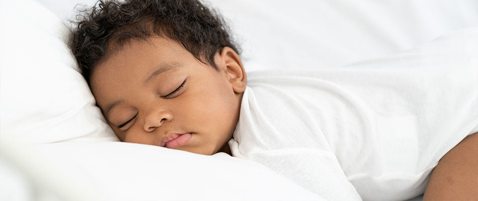 Ein Baby schläft friedlich auf einem weißen Bett, mit dem Kopf auf einem Kissen. Das Baby trägt ein weißes Oberteil und hat die Augen geschlossen.