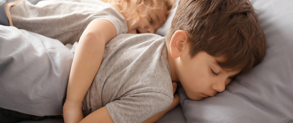 Zwei Kinder schlafen nebeneinander in einem Bett, beide tragen graue T-Shirts und sind in graue Bettwäsche gehüllt.