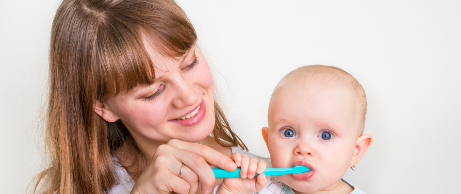 Eine Frau hilft einem Baby, die Zähne mit einer blauen Zahnbürste zu putzen. Die Frau lächelt, während das Baby neugierig in die Kamera schaut.