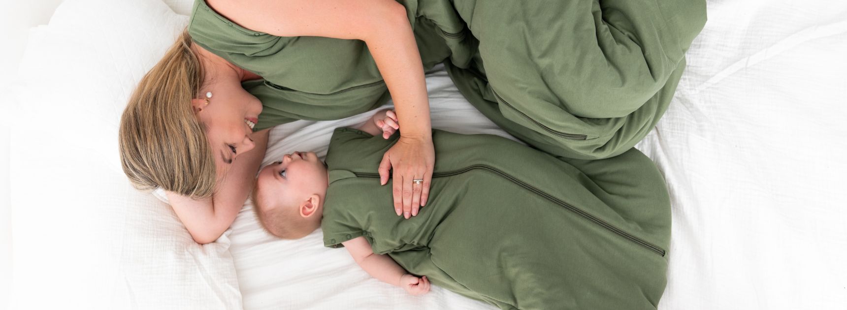 Eine Mutter liegt entspannt auf einem weißen Bett, gekleidet in einem grünen Schlafsack. Neben ihr liegt ihr Baby ebenfalls in einem passenden grünen Schlafsack. Beide schauen lächelnd.