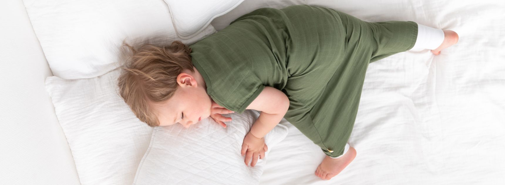 Ein kleines Kind schläft in einem grünen Schlafsack auf einem weißen Bett. Es liegt auf der Seite, mit einem Kissen unter dem Kopf.